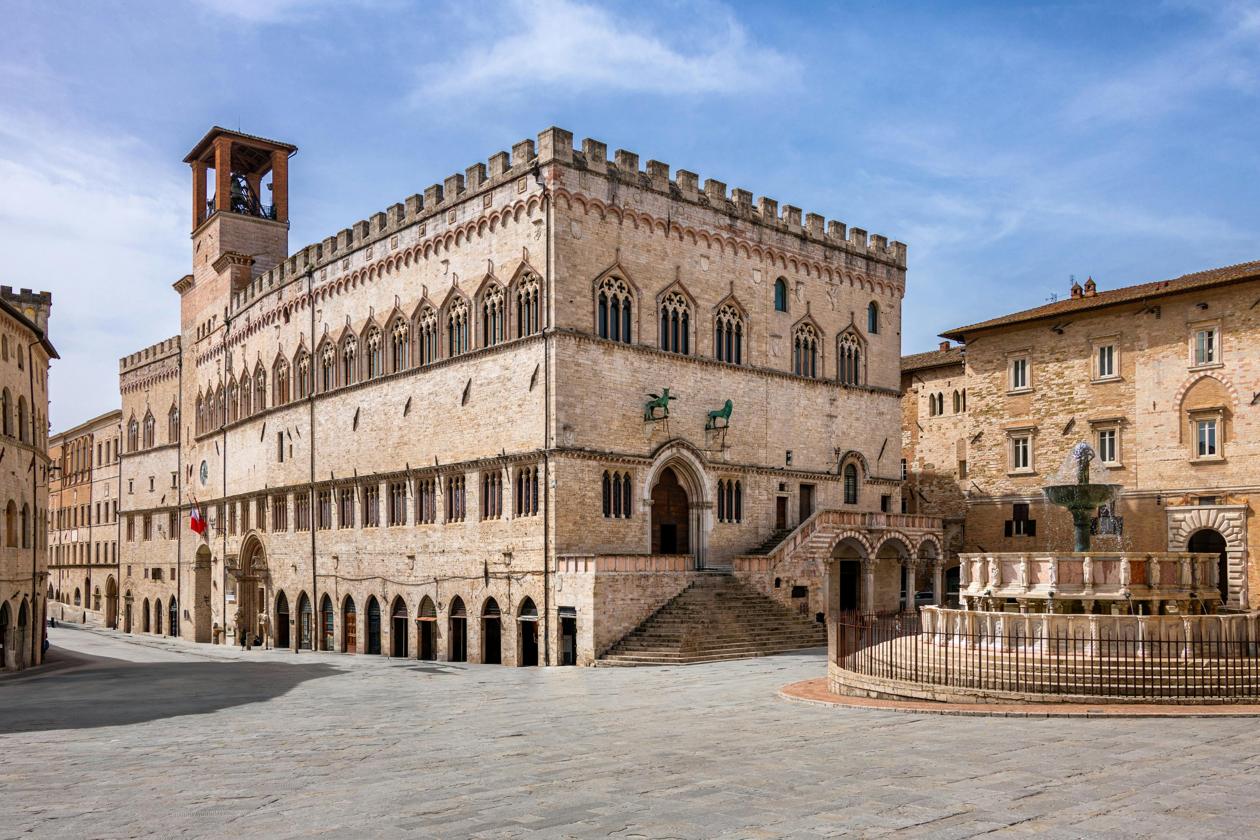 Edificio storico in pietra con finestre ad arco e scale, adiacente a una fontana in una piazza chiara e aperta sotto un cielo azzurro.
