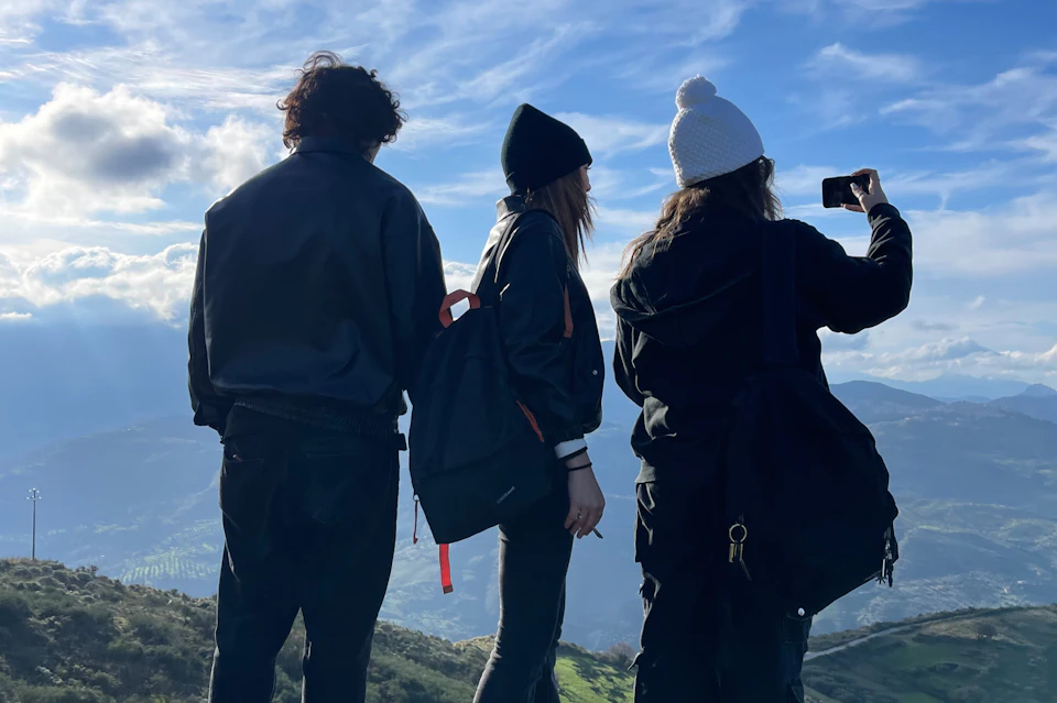 Three people in winter clothing stand on a hill, one taking a photo with a smartphone, with mountains and clouds in the background.