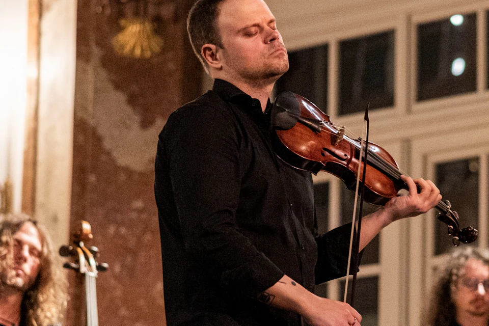 A violinist performs passionately in a classical music ensemble, with cellists and music stands visible in the background.