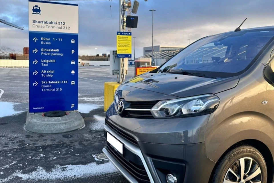 A gray van is parked in a lot near blue and yellow signs indicating directions for parking and terminals. Overcast sky in the background.