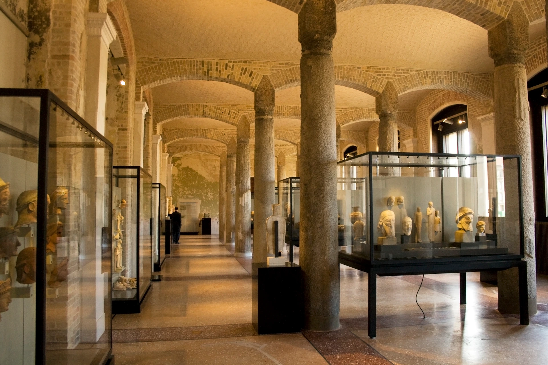 A museum hall with stone columns, arched ceilings, and glass display cases showcasing sculptures and artifacts.