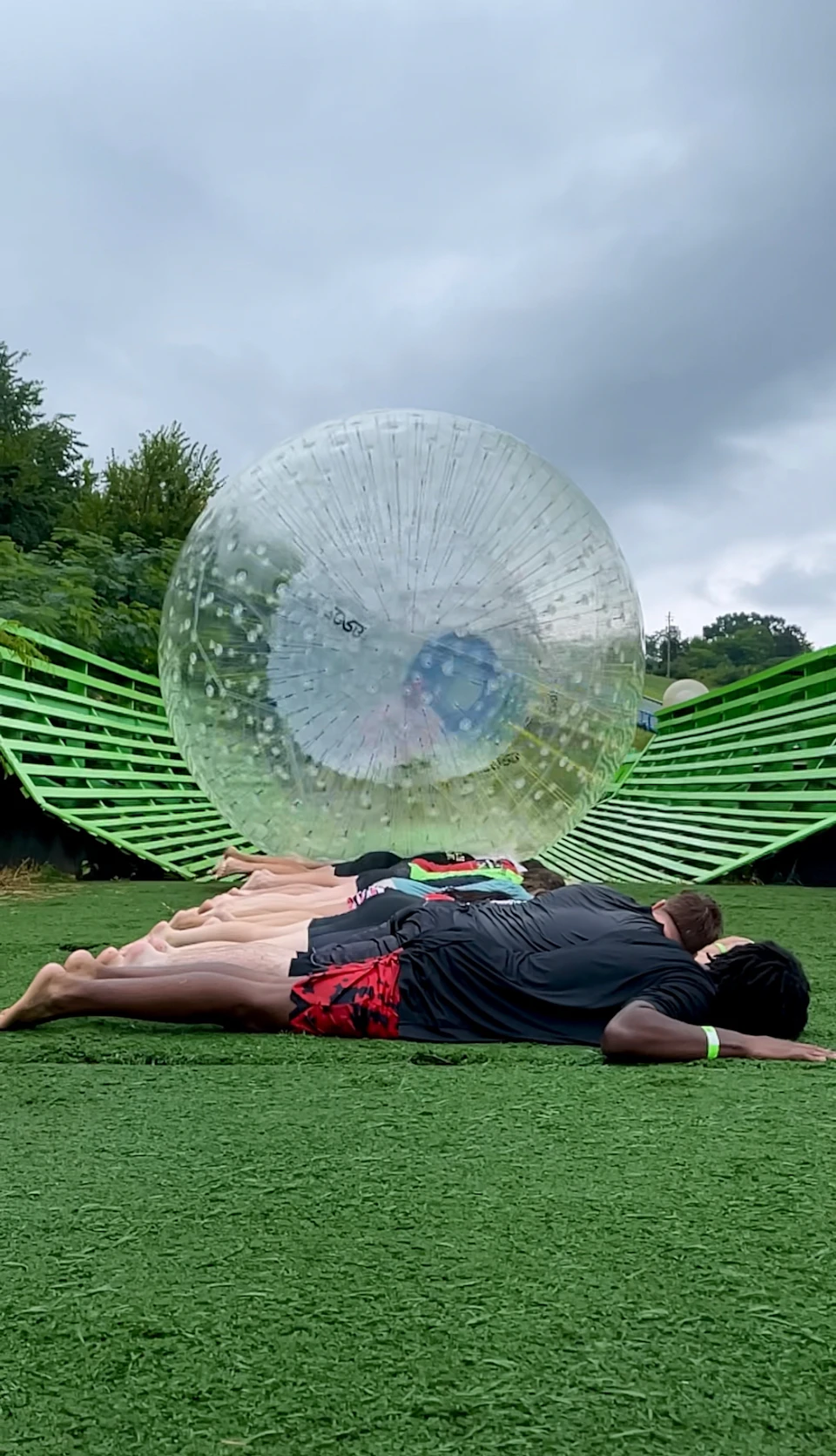 People lying in a row on green turf as a large inflatable ball rolls over them, with green structures on either side.