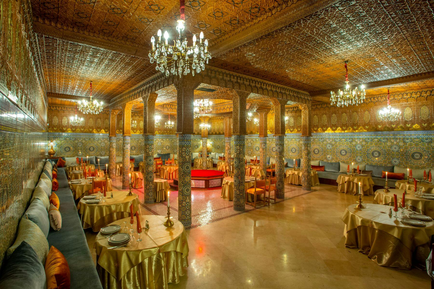 華やかな柱、シャンデリア、複雑な壁と天井のデザインが施されたエレガントなレストラン。 テーブルには金色のテーブルクロスが敷かれています。
