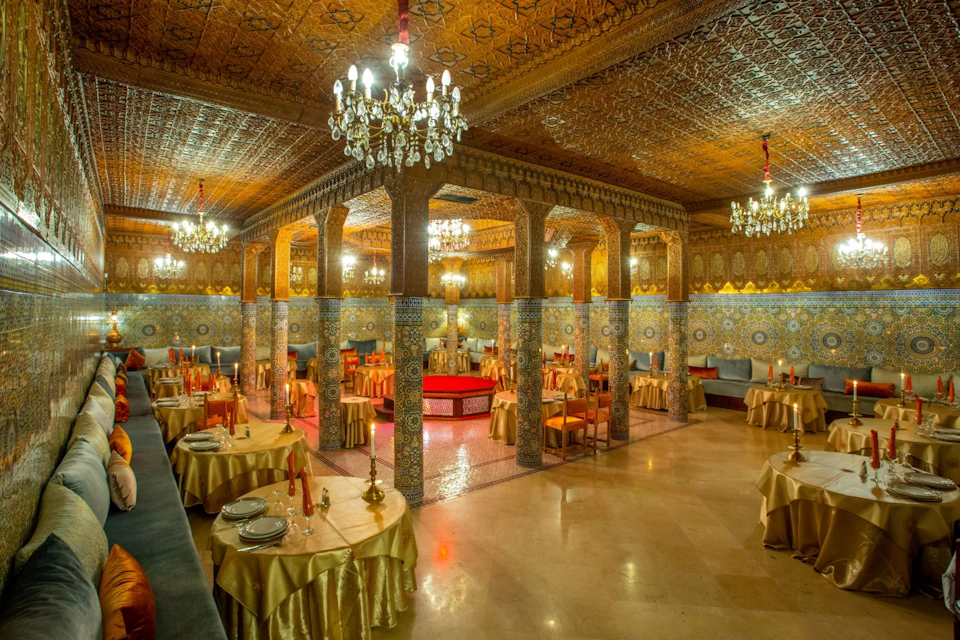 Elegant restaurant with ornate columns, chandeliers, and intricate wall and ceiling designs. Tables are set with golden tablecloths.