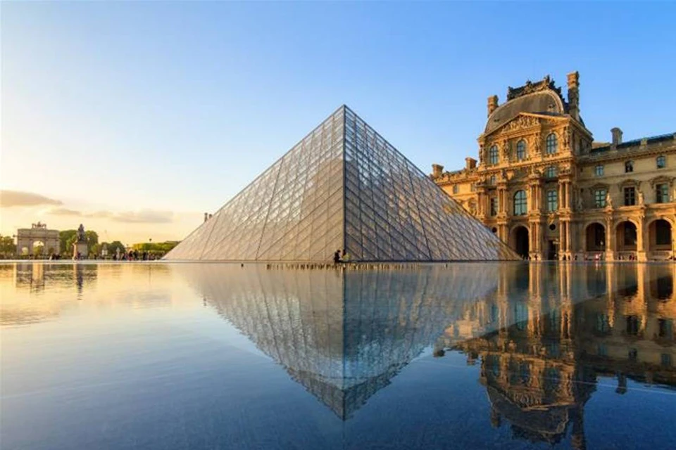 A glass pyramid with a reflection in water in front of a historic building under a clear sky at sunset.