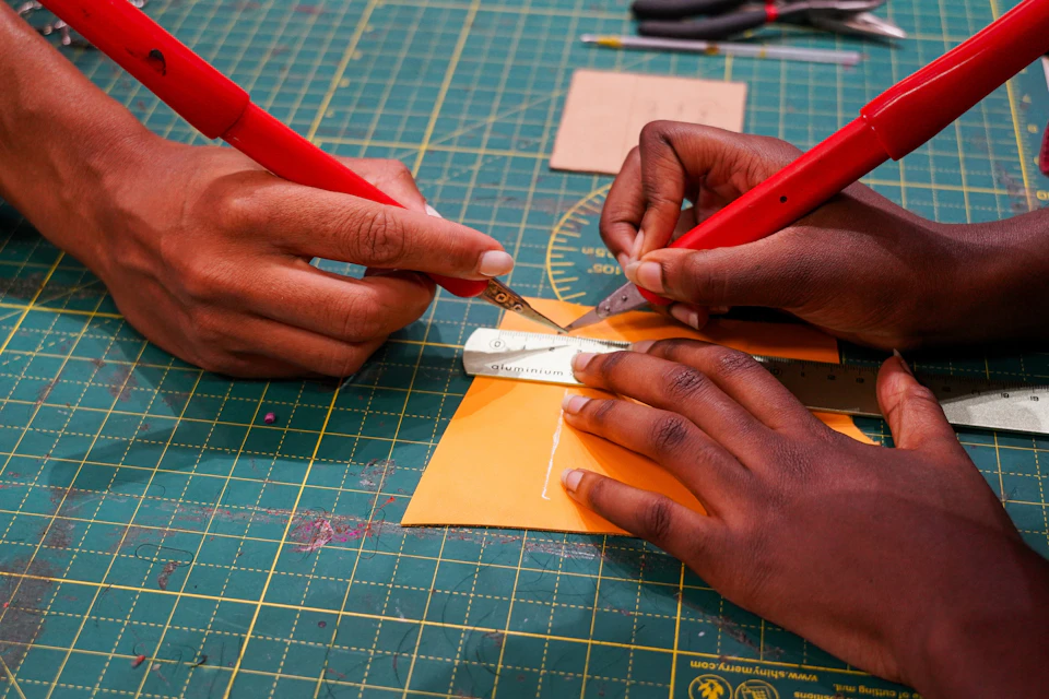 Two hands using red craft knives to cut an orange paper sheet on a green cutting mat. Rulers and a white eraser are visible.