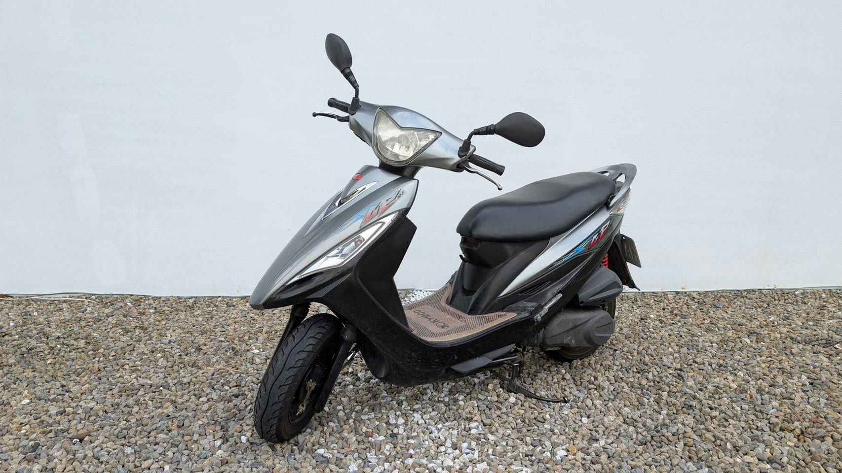 A silver and black scooter parked on gravel against a plain white wall.