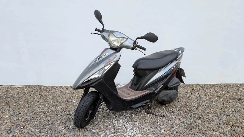 A silver and black scooter parked on gravel against a plain white wall.