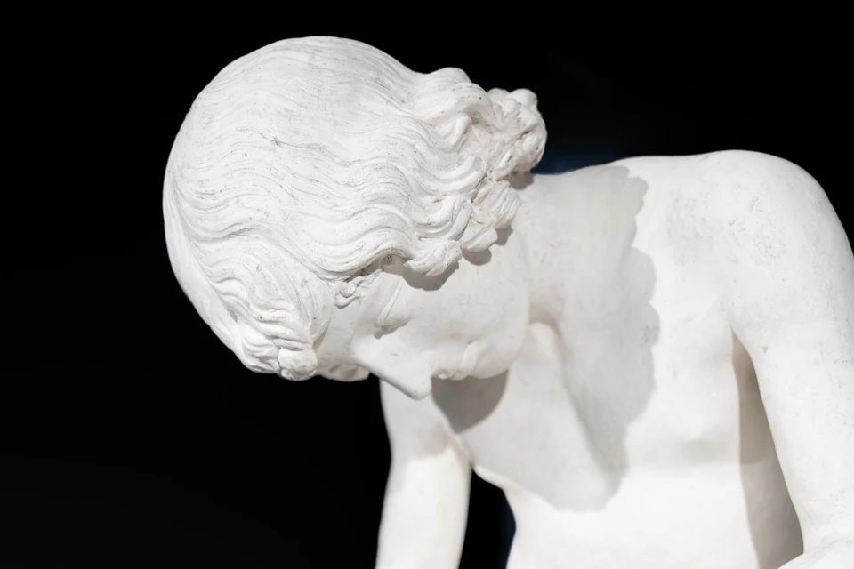 Close-up of a white marble statue depicting a figure with curly hair and a downward gaze against a dark background.