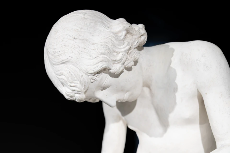 Close-up of a white marble statue depicting a figure with curly hair and a downward gaze against a dark background.