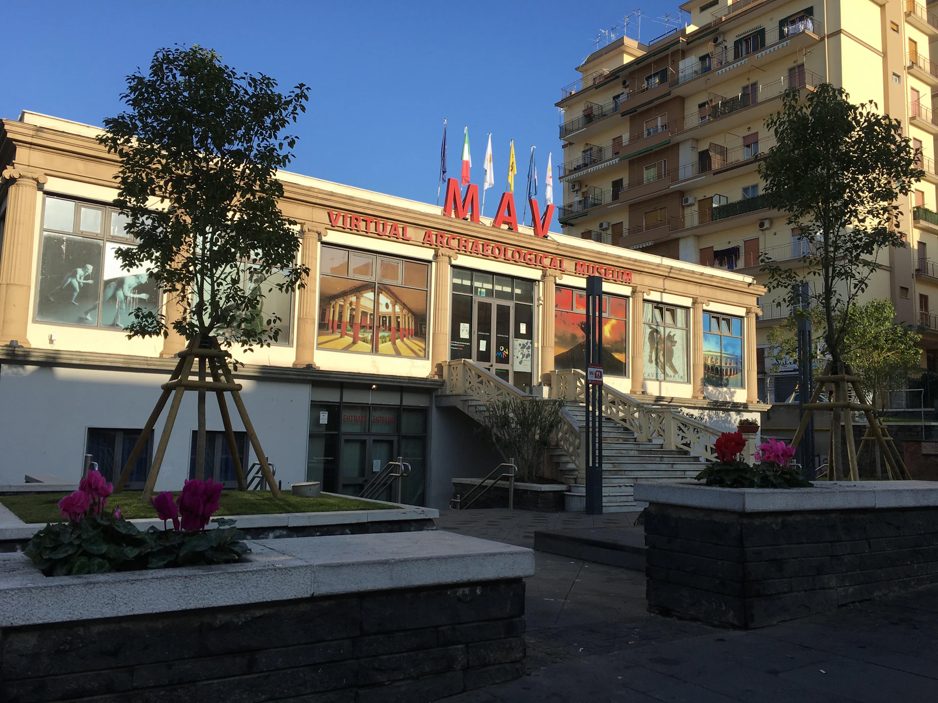 Building with a "MAV Virtual Archaeological Museum" sign, colorful murals, and a staircase in front. Residential building in the background.
