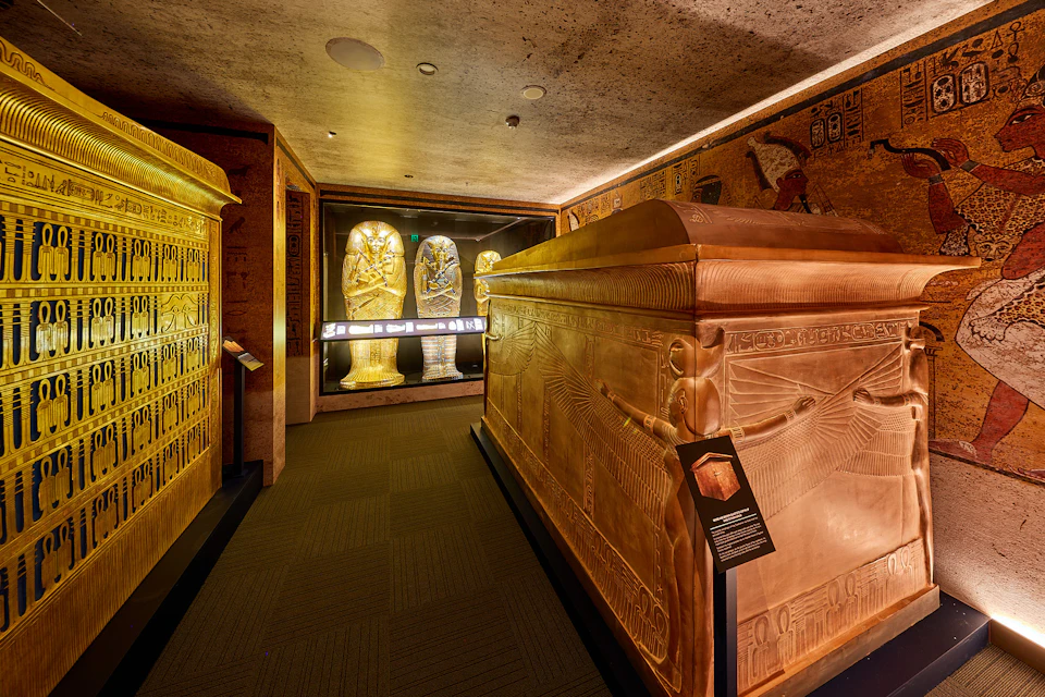 A museum exhibit featuring an Egyptian sarcophagus, hieroglyphic-inscribed walls, and two illuminated statues in the background.