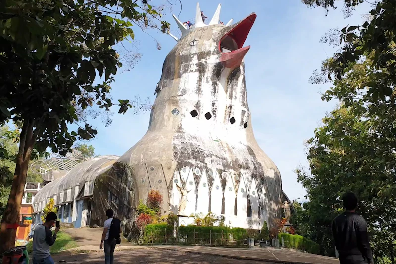 A large building shaped like a chicken with a red beak, surrounded by greenery and a person walking nearby.