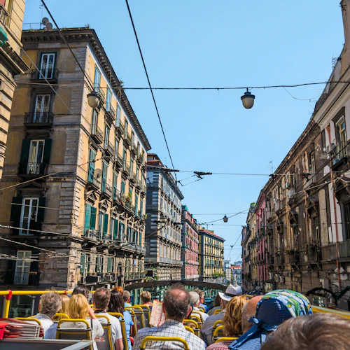 En grupp människor på en dubbeldäckad busstur på en smal stadsgata kantad av höga, historiska byggnader under en klar himmel.