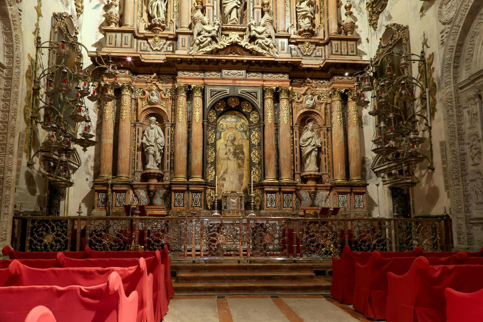Ozdobny ołtarz kościelny z posągami, misternymi rzeźbami i krzesłami pokrytymi czerwienią na pierwszym planie.