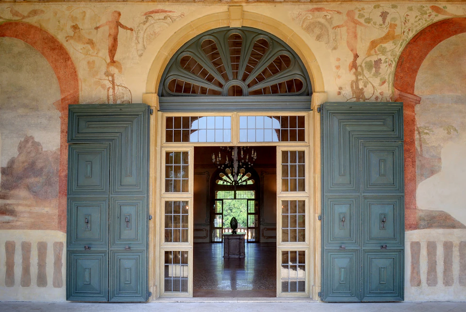 Ornate double doorway with large turquoise doors open to a grand hall showcasing a chandelier and a distant statue.
