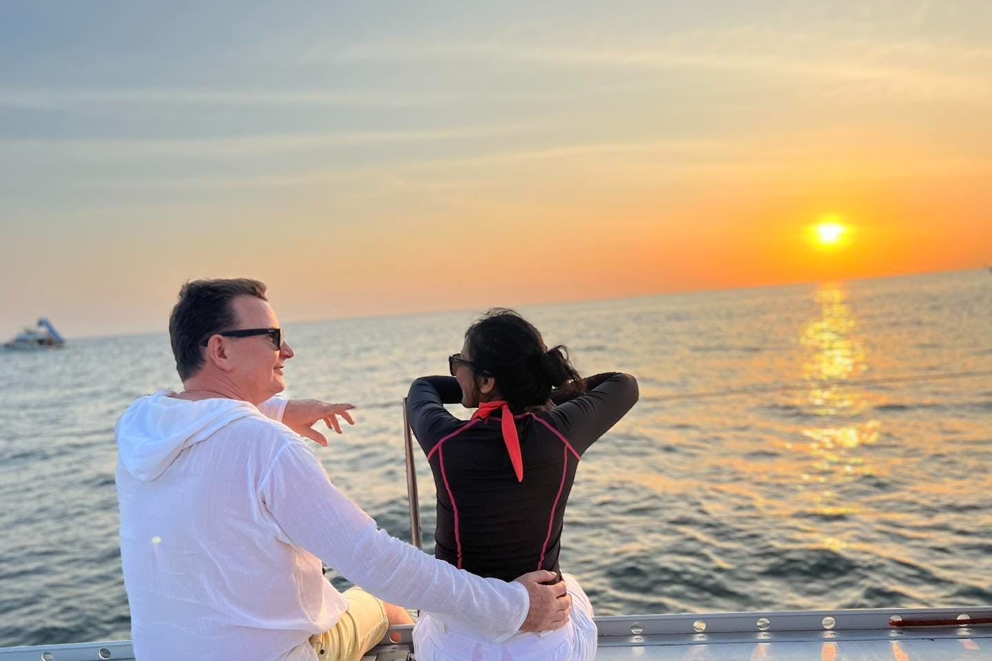 Ένας άντρας και μια γυναίκα κάθονται σε μια βάρκα στο ηλιοβασίλεμα, με τα χέρια της γυναίκας να στηρίζονται σε μια ράγα και μια μακρινή βάρκα να φαίνεται στον ορίζοντα.