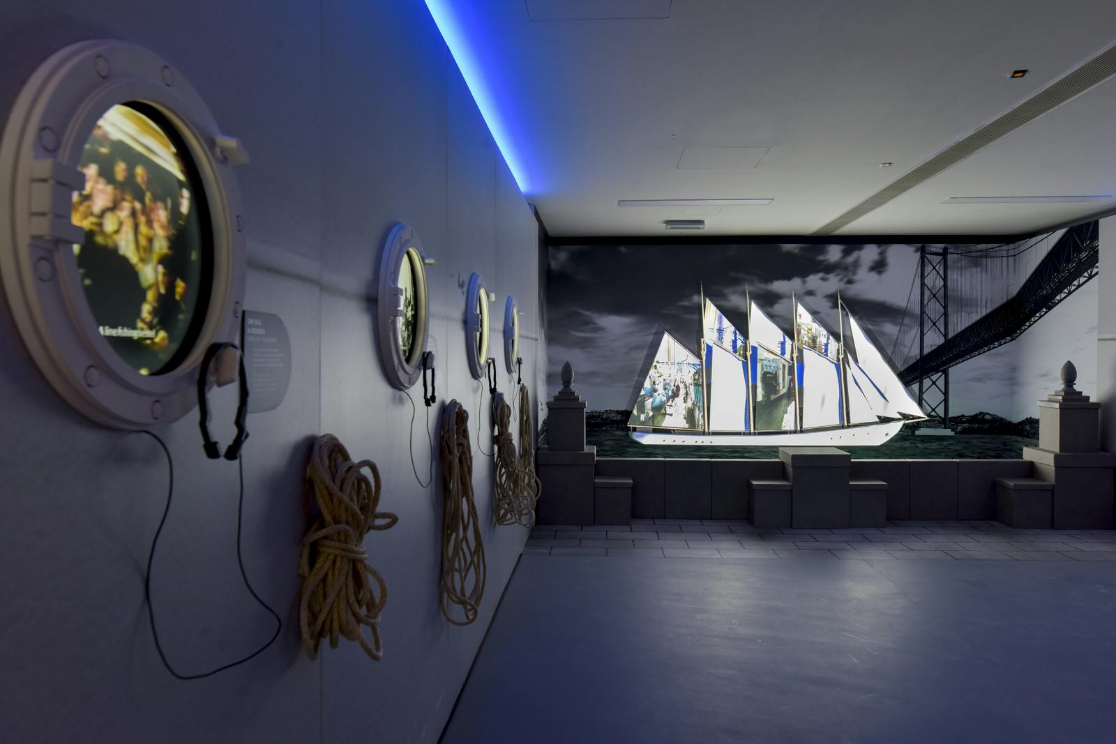 A museum exhibit with nautical ropes on the wall and a large screen displaying a sailboat at sea.