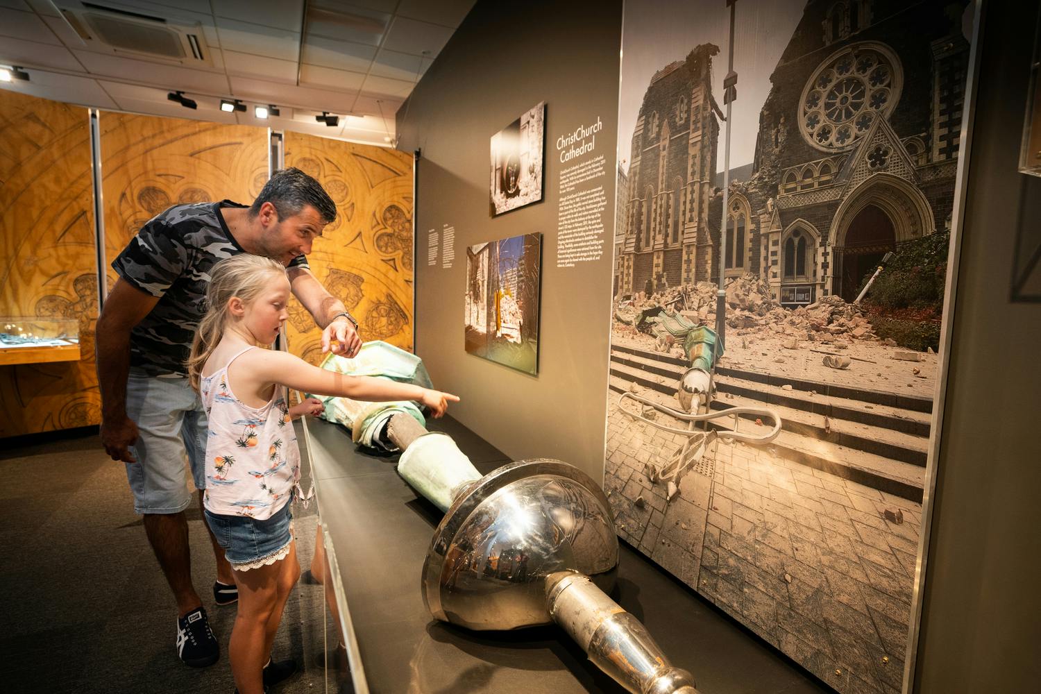 A man and a girl observe a display in a museum, featuring a large metallic object and historical photographs on the wall.