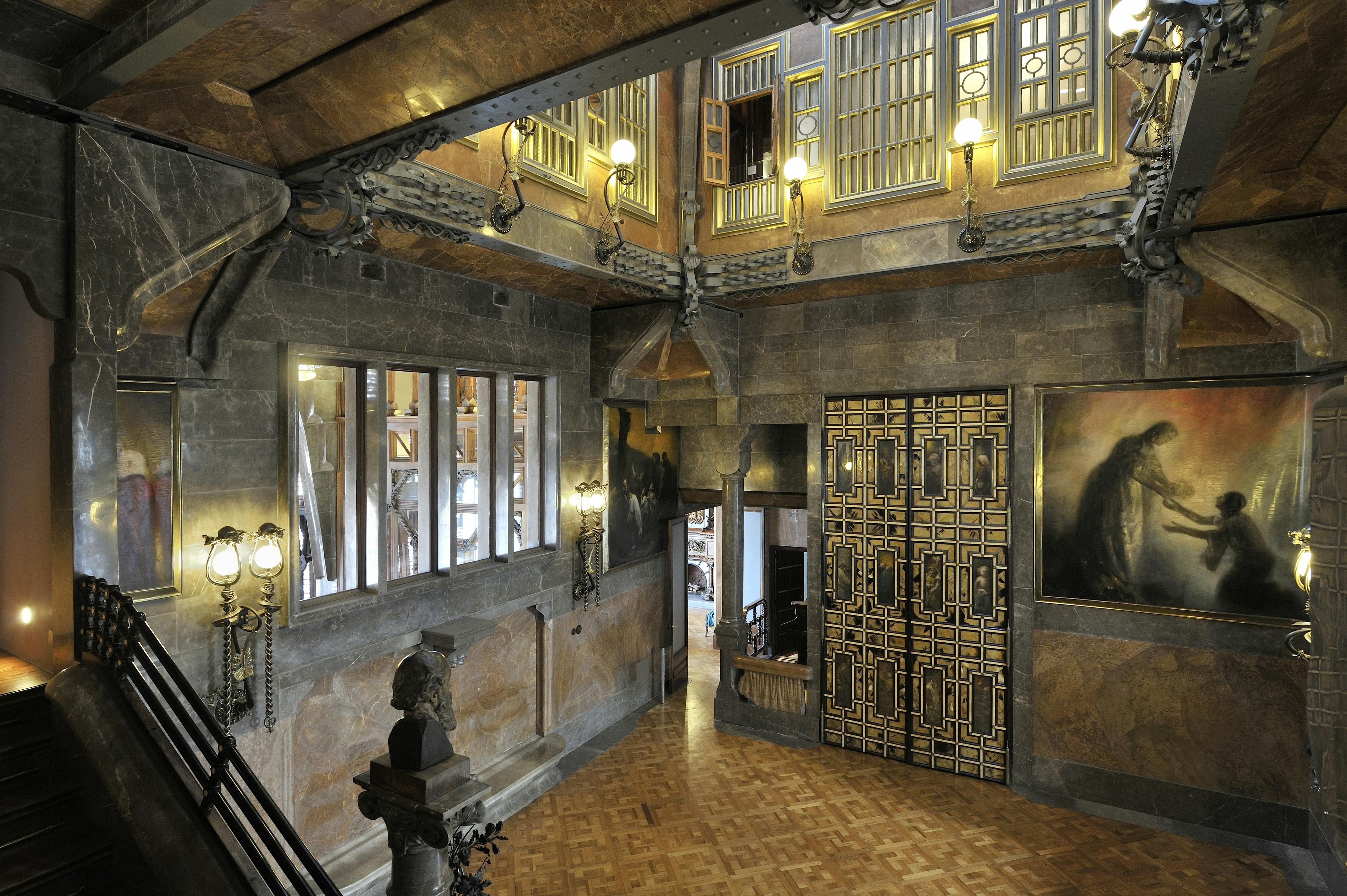Ornate interior with wood and stone details, ironwork, large decorative door, bust statue, and balcony with lamps.