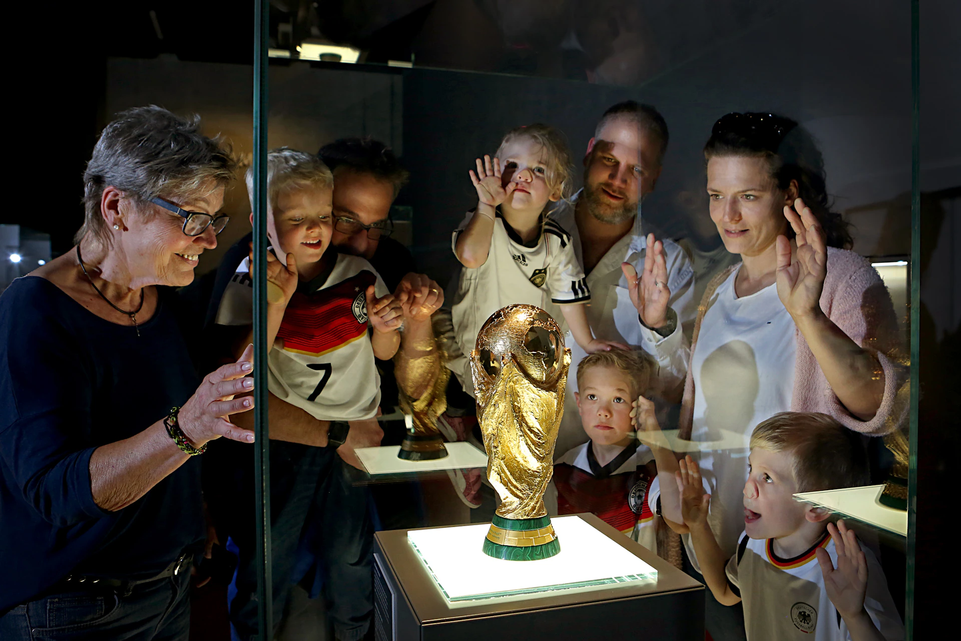 Several people, including children wearing soccer jerseys, look at a lit World Cup trophy inside a glass display case.