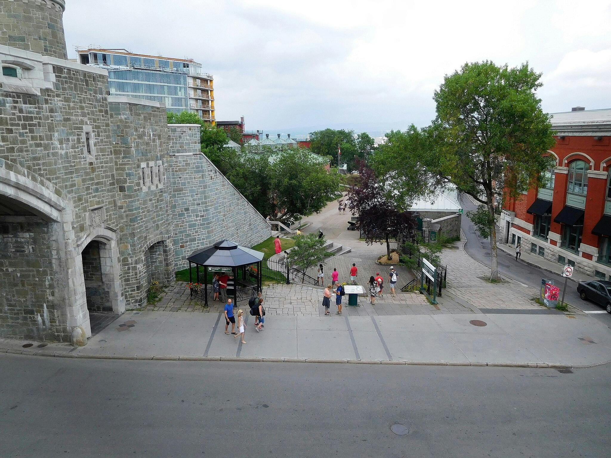 通りに隣接する石造りの建物で、数人が歩道や木々、周辺の建物を歩いています。