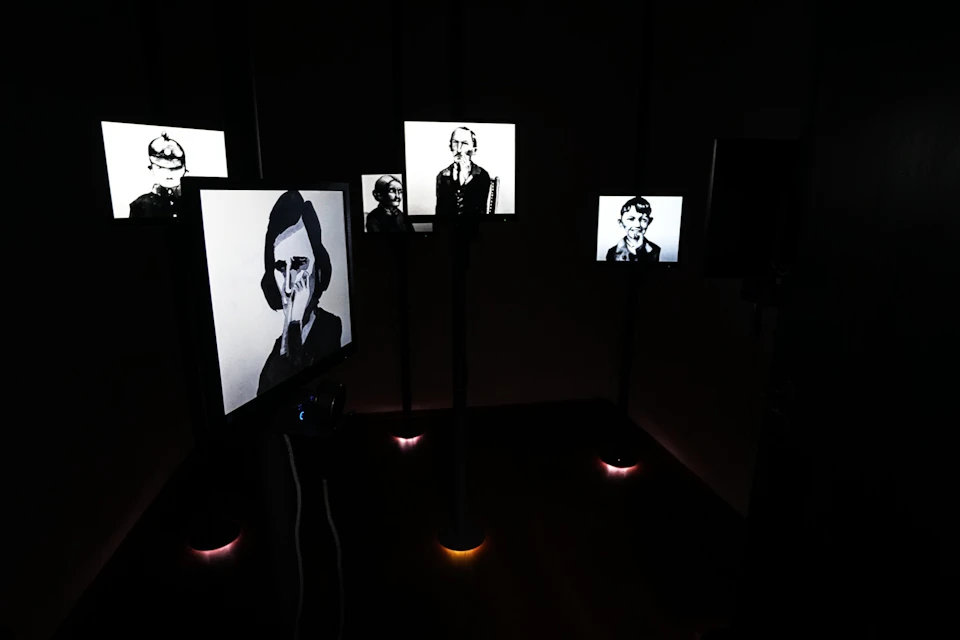 Dark room with several lit screens displaying black-and-white portraits of people.