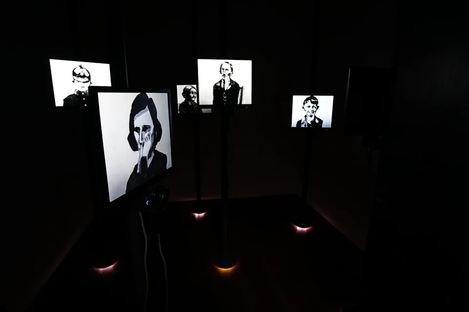 Dark room with several lit screens displaying black-and-white portraits of people.
