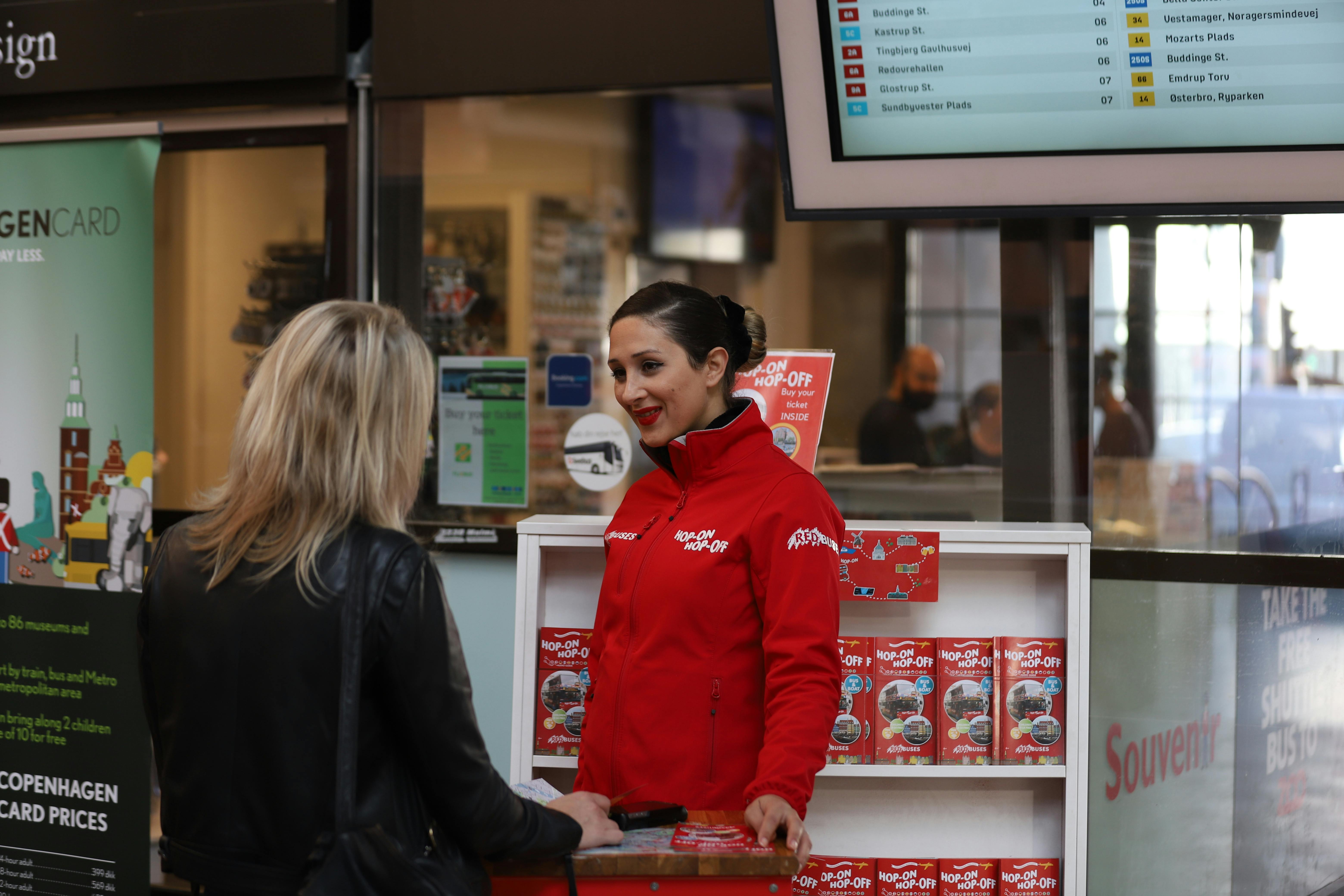 A woman in a red jacket, labeled "Hop On Hop Off," stands behind a counter talking to another woman in a black jacket.