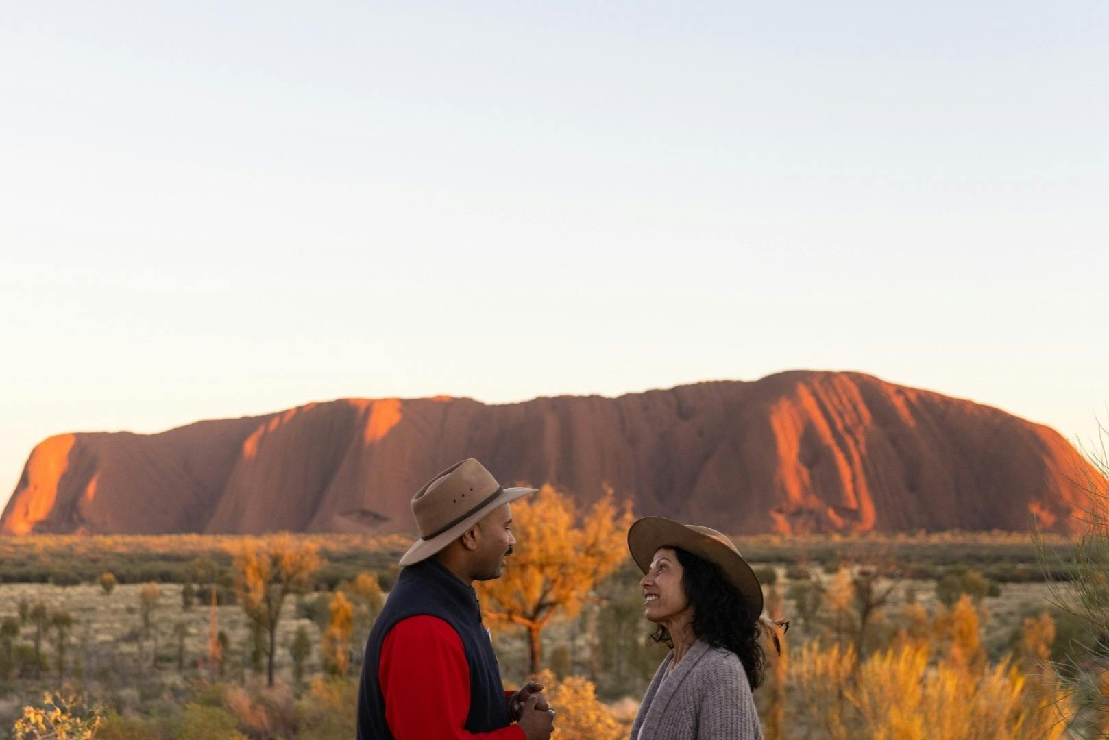 Dues persones amb barrets conversen davant d'una gran formació rocosa en un paisatge desèrtic al capvespre.
