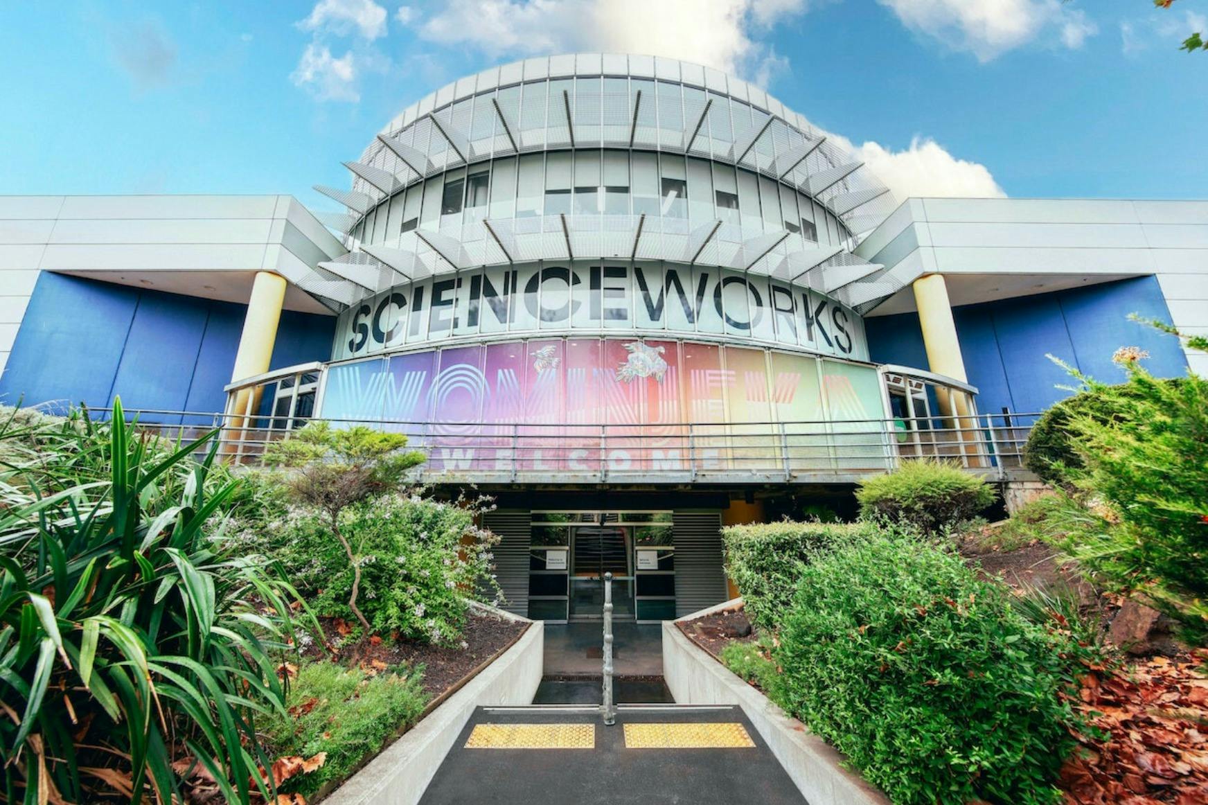 A entrada para um edifício moderno com a designação "Scienceworks", rodeado de vegetação e com uma fachada de vidro colorido.