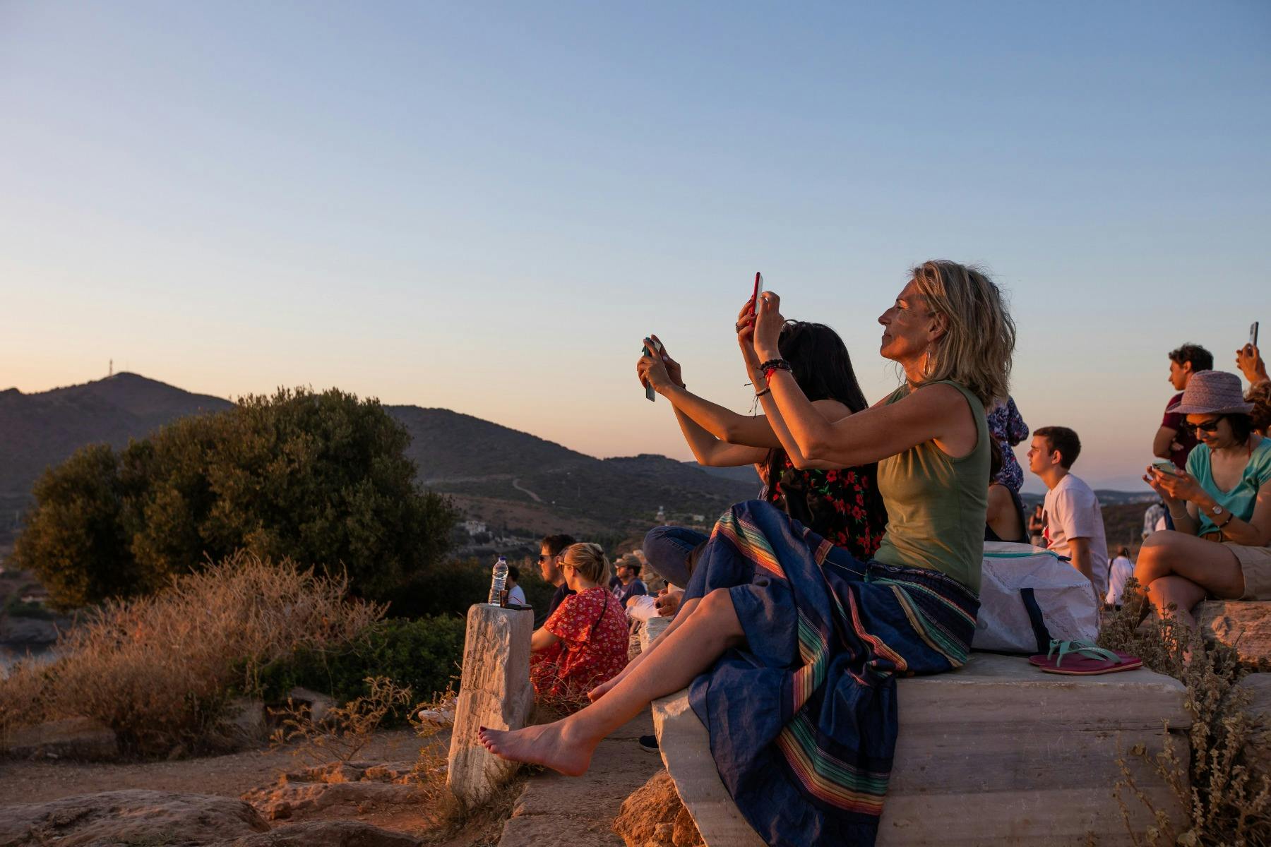 Travelers sitting on ancient stones, photographing the sunset over the Aegean from Cape Sounion.