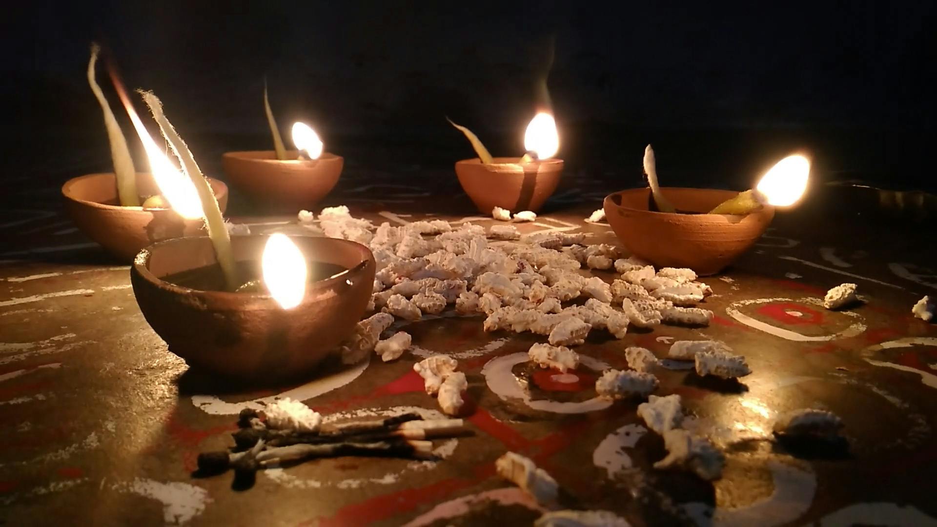 Vier Öllampen aus Ton mit brennenden Dochten auf einer verzierten Oberfläche, umgeben von weißen, strukturierten Stücken.