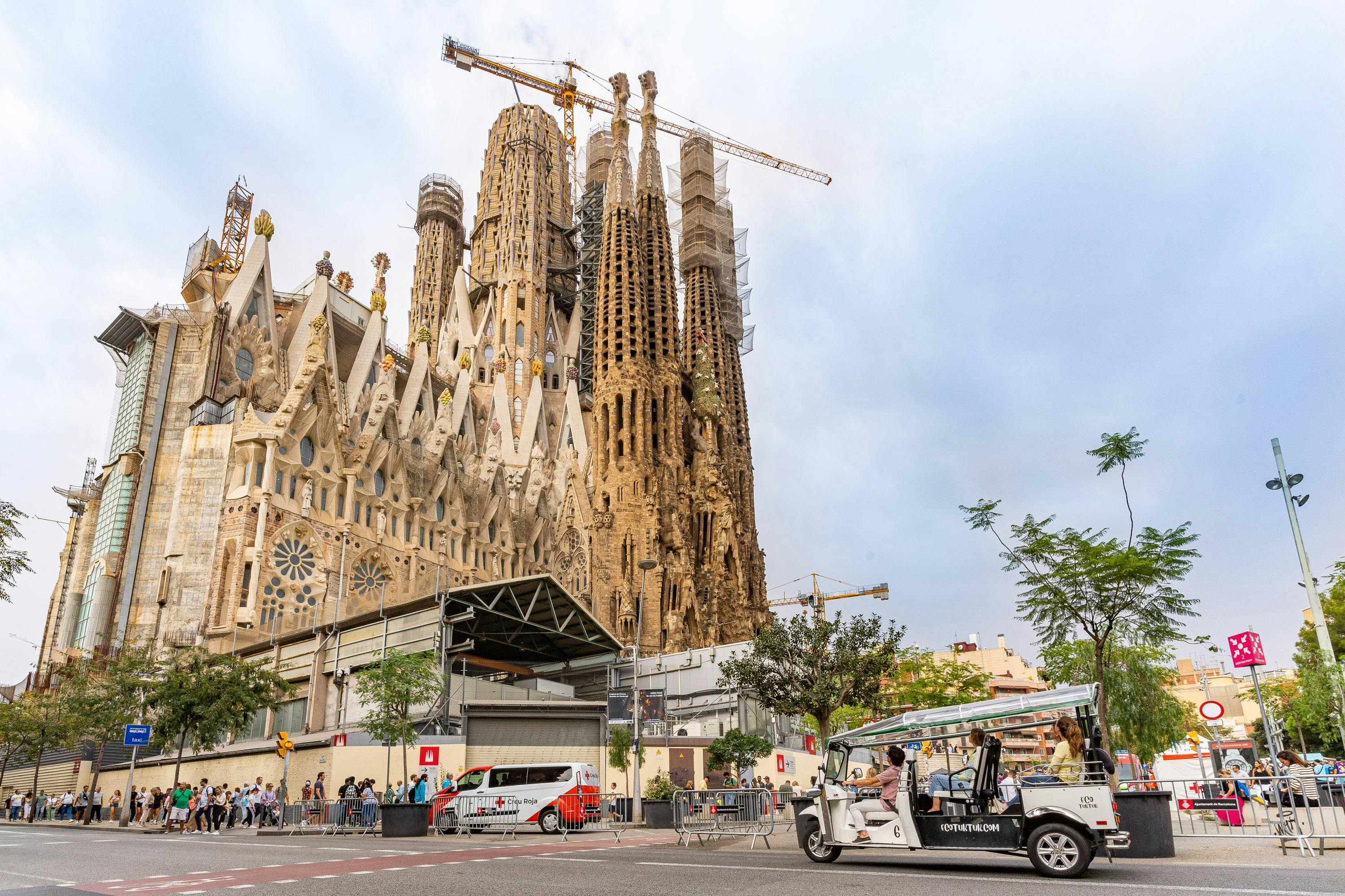 En stor, invecklad katedral under uppbyggnad med kranar, omgiven av turister och ett förbipasserande vitt elfordon.