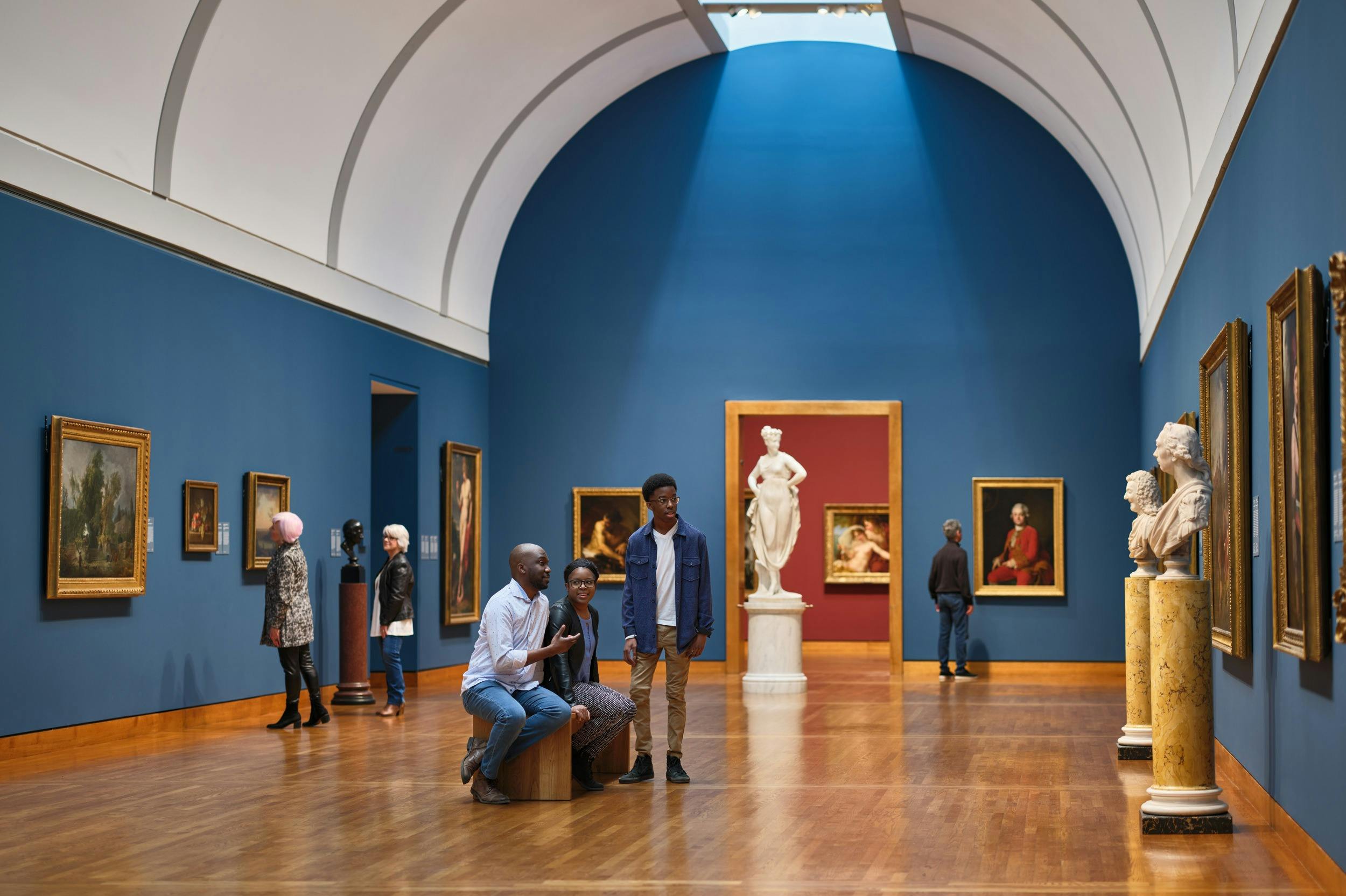 People observing paintings and sculptures in a spacious art gallery with blue walls and wooden flooring.