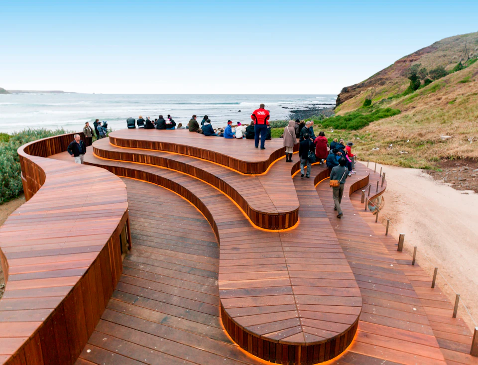 People sitting on tiered wooden seating overlooking a coastal view with grassy hills and a blue sky in the background.