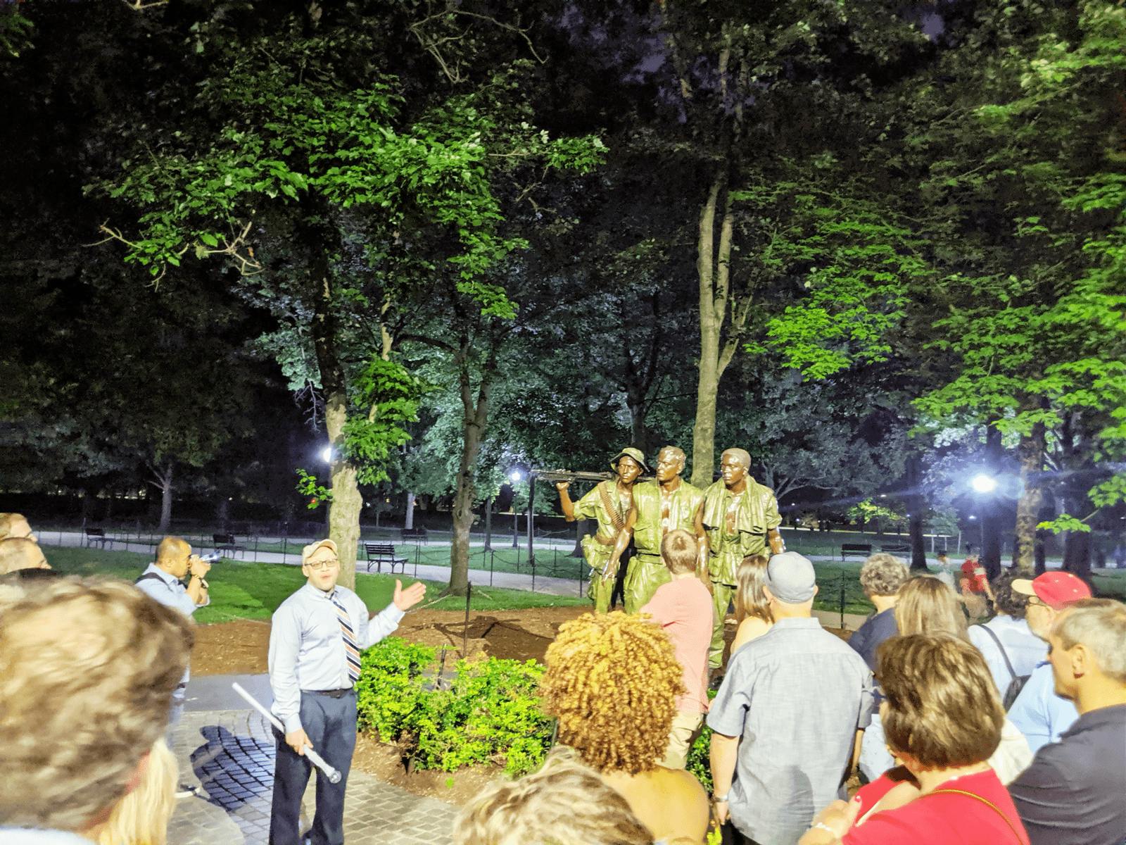 한 무리의 사람들이 밤에 공원에서 네 명의 군인 동상 앞에서 말하는 한 남자의 말을 듣고 있습니다.