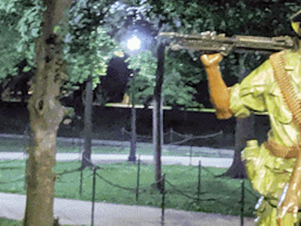 A group of people listens to a man speaking in front of a statue of four soldiers in a park at night.