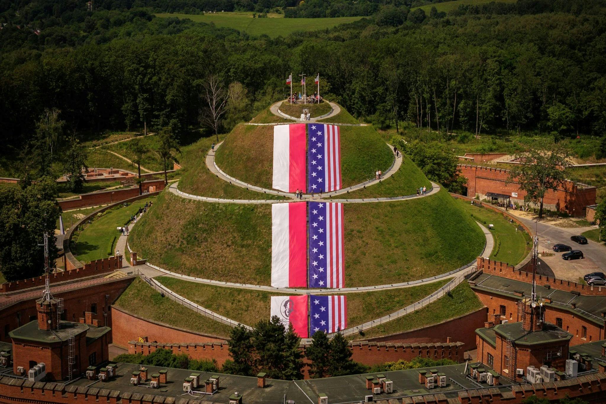 A grassy mound with large flags displayed on its surface, surrounded by pathways and trees in the background.
