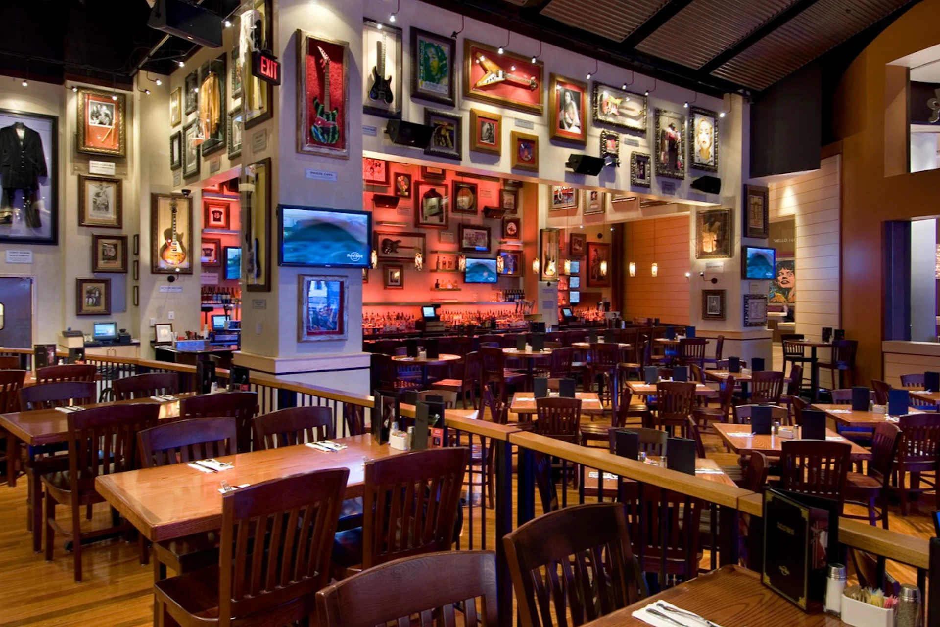 Restaurant interior with wooden tables and chairs, framed memorabilia on walls, TVs, and a bar area in the background.