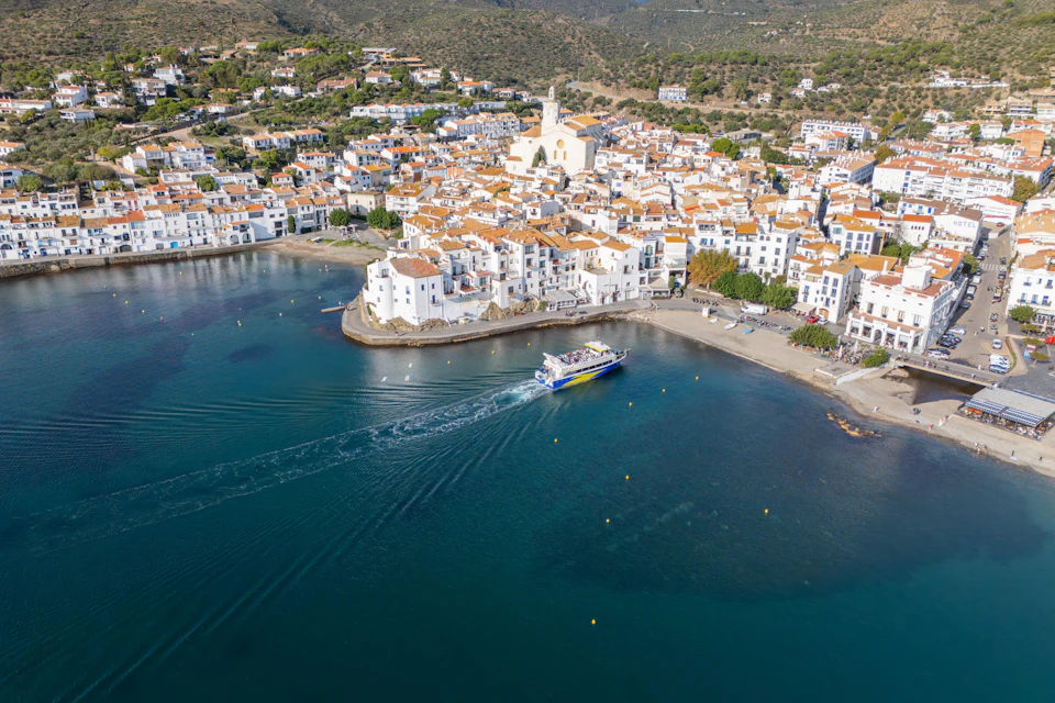 A coastal town with white buildings and red roofs lies on either side of a bay, with a large boat gliding through the water.