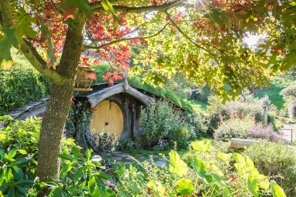A small hobbit-style house with a round wooden door surrounded by lush greenery and colorful flowers under sunlight.