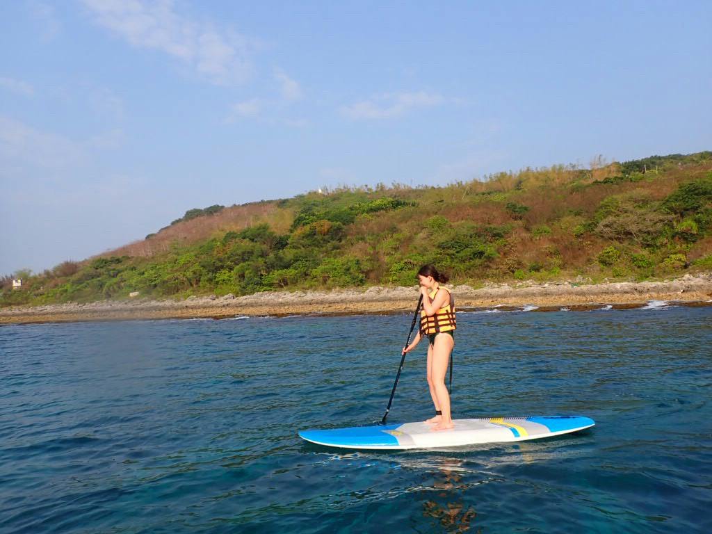 A person paddleboarding on calm blue water near a lush, tree-covered shoreline under a clear sky.