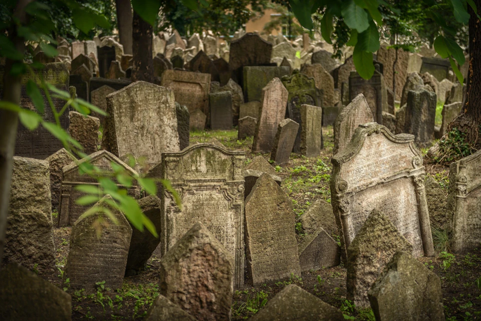 Several old, weathered gravestones closely packed in a cemetery, surrounded by greenery and trees.