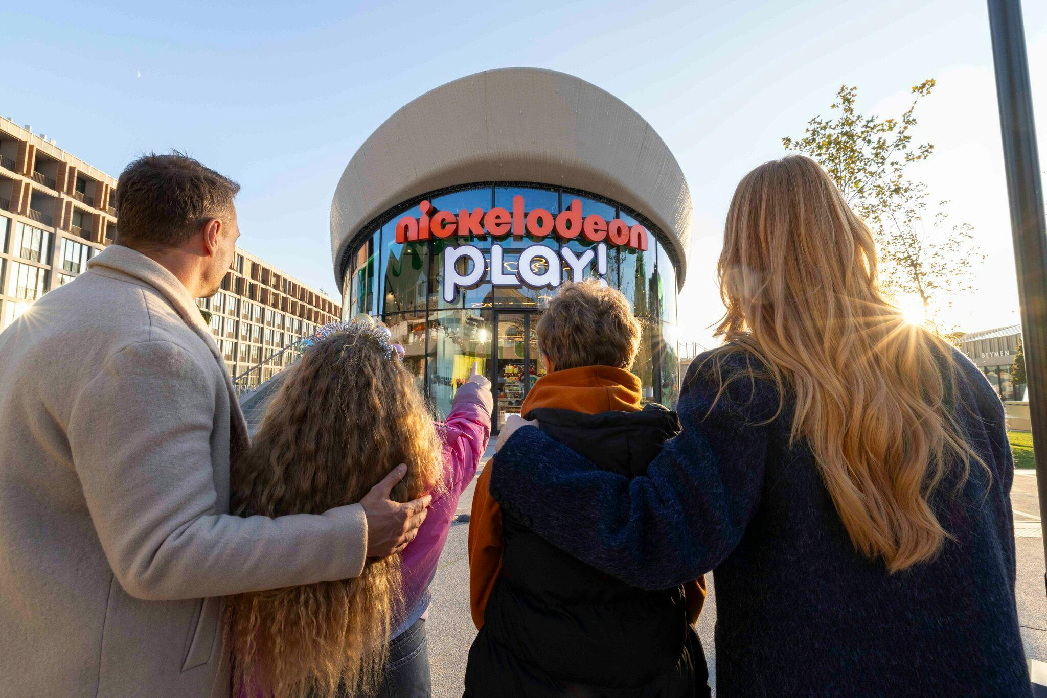 Een groep van vier mensen staat buiten en wijst naar een gebouw met een bord waarop 'Nickelodeon Play' staat.
