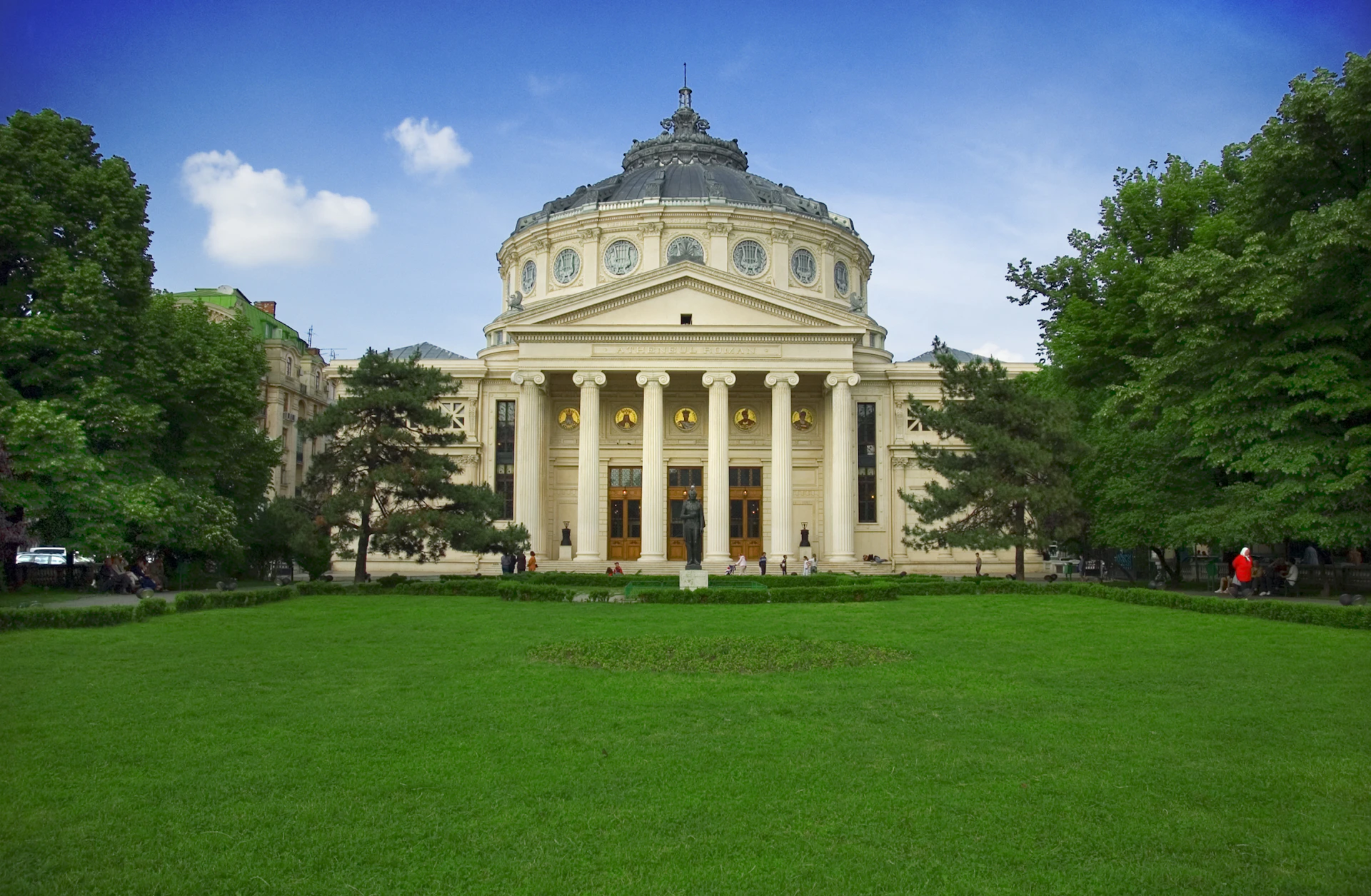 Grand building with a domed roof and columns, set in a park with green lawn and trees; clear blue sky in the background.