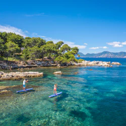 Två personer paddlar i en klar, blå kustvik med klippiga stränder och frodiga gröna träd under en klarblå himmel.