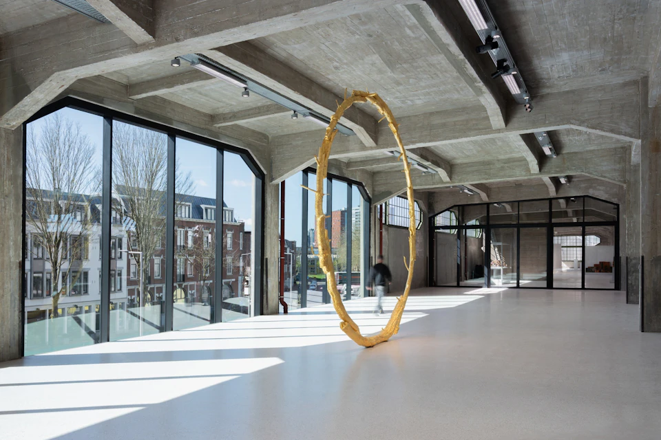 A modern art gallery with large windows, showcasing a circular golden sculpture in a spacious, light-filled room.