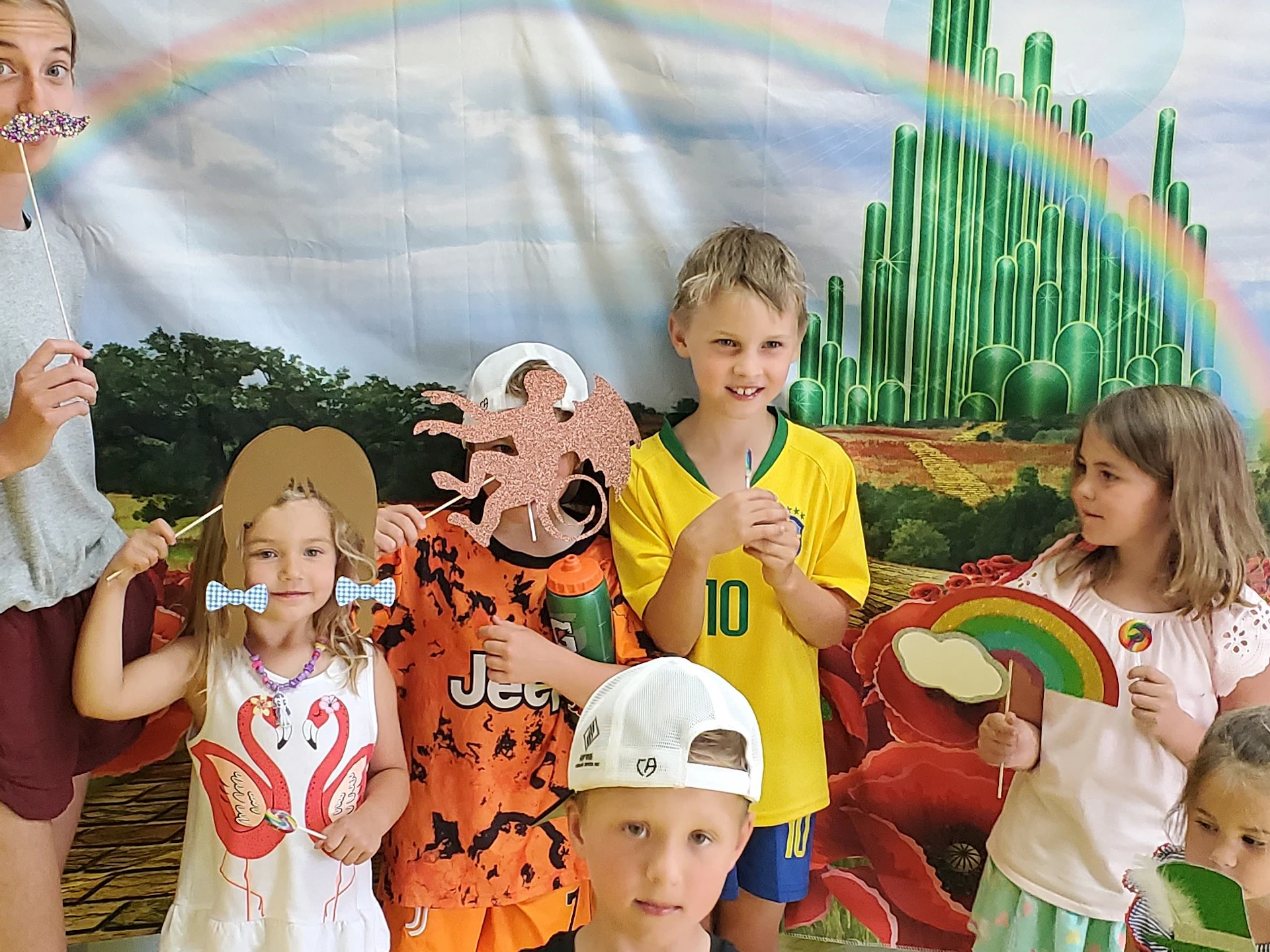 Six children pose with props in front of an Oz-themed backdrop, featuring a rainbow and the Emerald City.