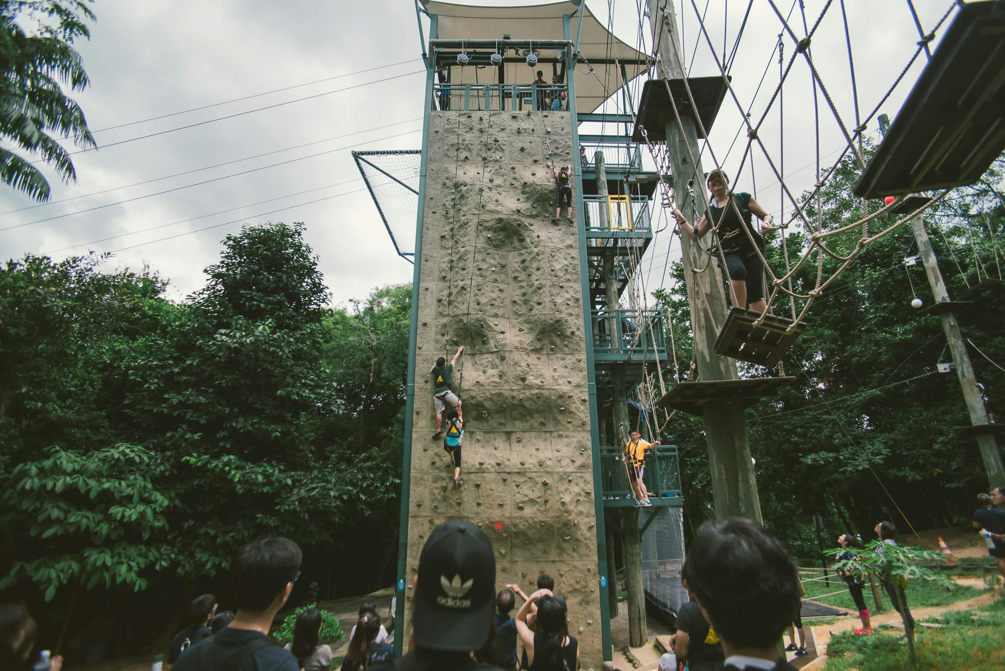 People watching two climbers on a tall outdoor climbing wall, while another person navigates a rope course beside it.